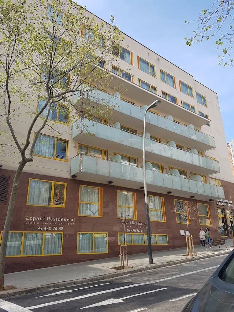 La fachada de un edificio moderno de varios pisos en tonos beige y marrón, con balcones de cristal y el letrero "Lepant Residencial" visible en la planta baja. Hay árboles jóvenes y una calle con marcas de paso de peatones en frente.
