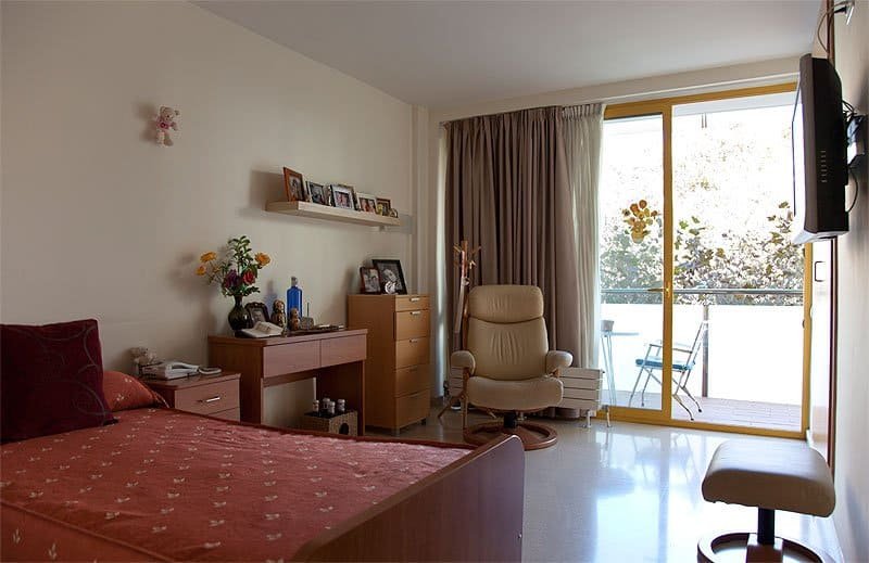 Una habitación individual luminosa en una residencia, con una cama con colcha estampada, un escritorio de madera con objetos personales, un sillón reclinable de cuero claro y acceso a un balcón soleado.