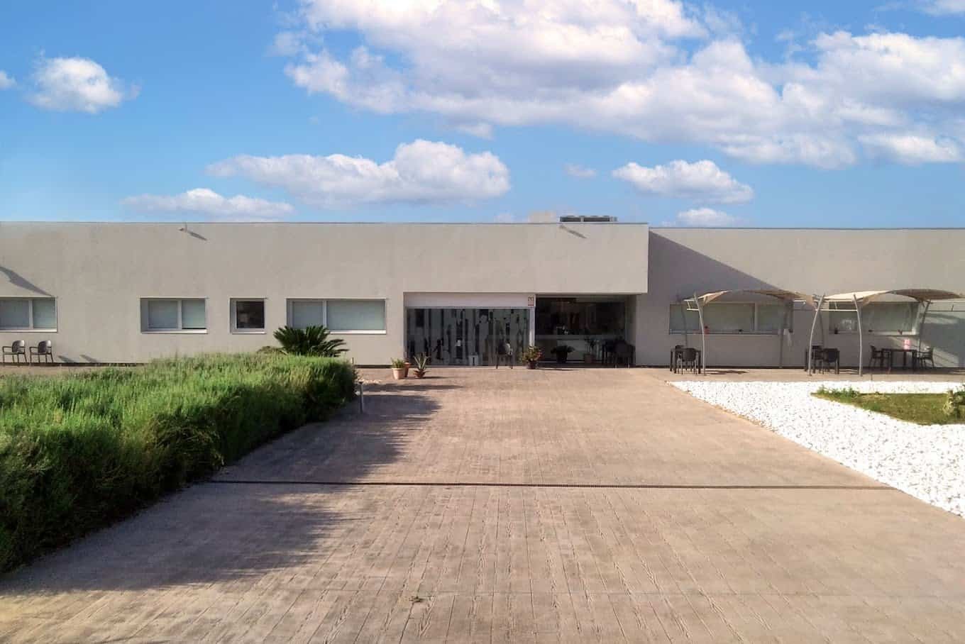Vista exterior de un edificio de una sola planta en un día soleado. El edificio es de color blanco y tiene grandes ventanales. Un camino pavimentado conduce a la entrada, flanqueado por una zona de arbustos bajos y un área de grava blanca con mesas y sombrillas.