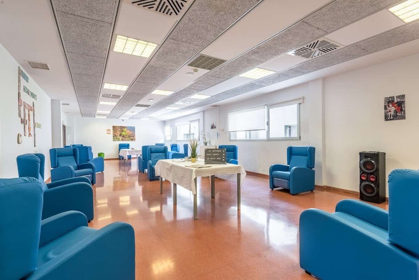 Amplio salón con suelo de terrazo, paredes blancas y techo con paneles acústicos. Está amueblado con sillones reclinables de color azul brillante dispuestos en semicírculo. En el centro hay una mesa de actividades cubierta con un mantel blanco y un letrero que dice "Toca Creixer". Un altavoz y varias fotos cuelgan en las paredes.