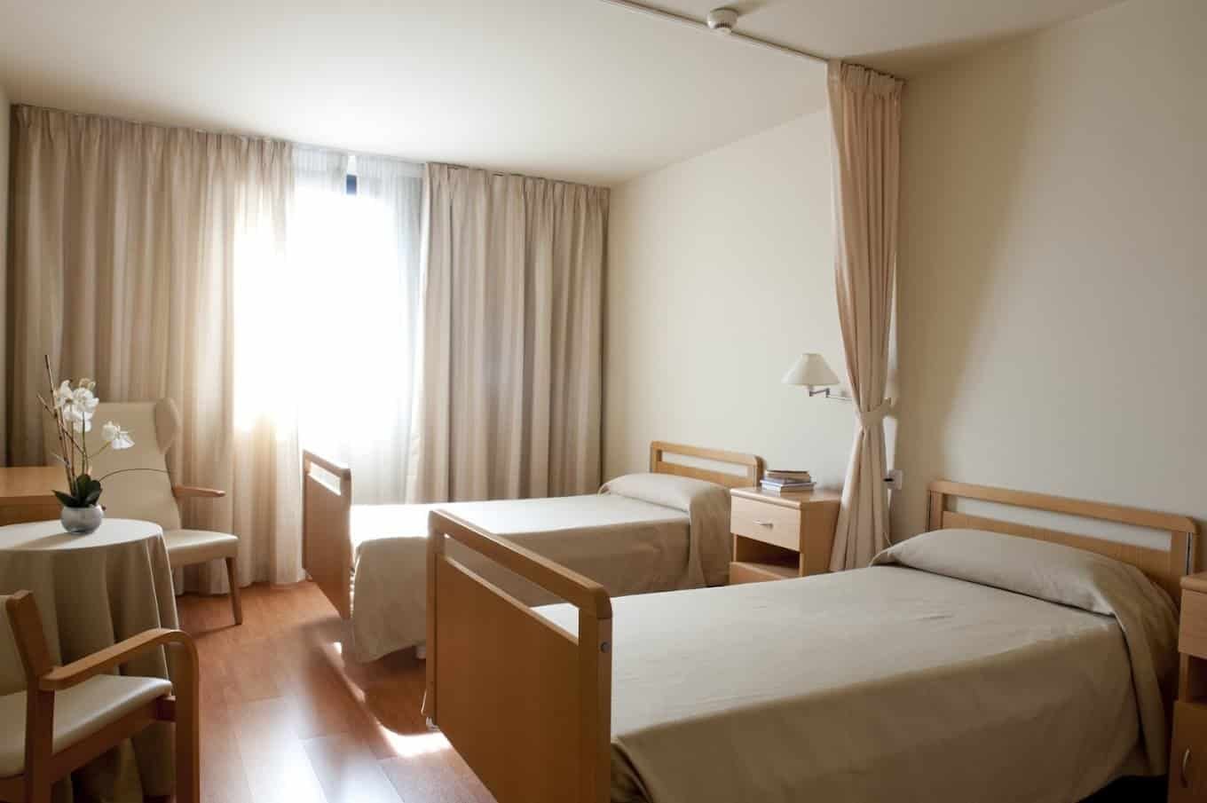 Habitación doble de una residencia con dos camas individuales, paredes claras, suelo de madera y grandes cortinas beige que cubren una ventana. Hay una mesita de noche, una silla y una orquídea blanca.