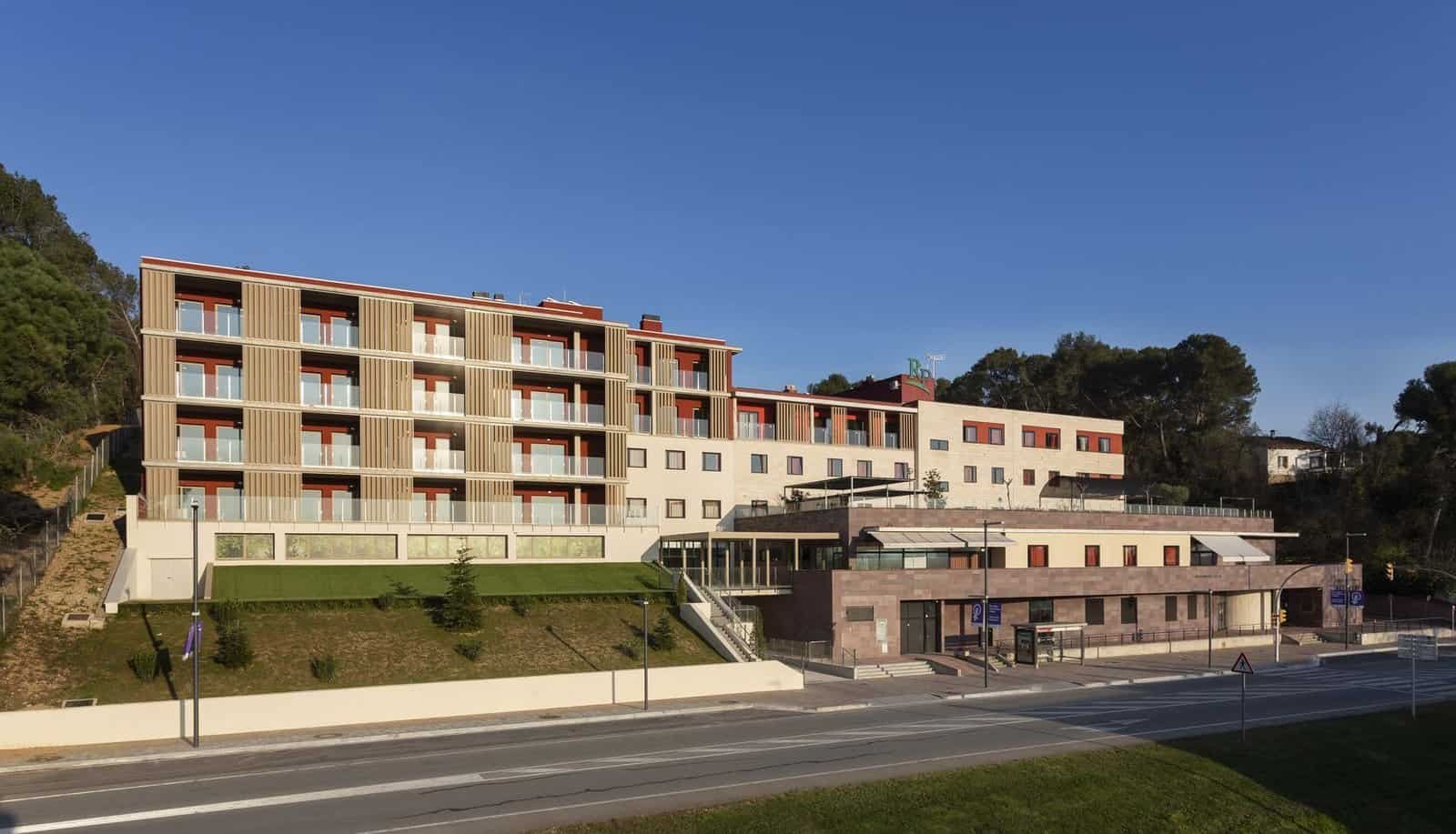 Vista exterior de un edificio de varios pisos en una zona de montaña. El edificio combina fachadas de color rojo con balcones acristalados y una base de piedra oscura.