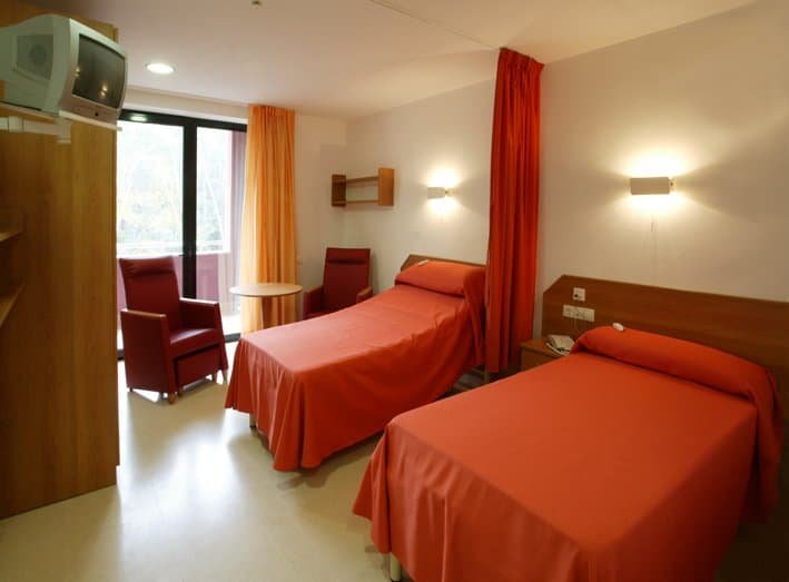 Una habitación con dos camas individuales, dos sillones rojos y una mesa redonda pequeña. La habitación está iluminada con luz natural que entra por la ventana.