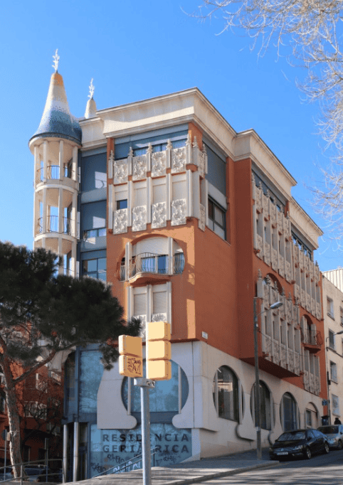Un edificio de estilo modernista con una fachada de colores cálidos, una torre distintiva con un tejado azul y balcones con barandillas decorativas. En la planta baja se aprecia un letrero que dice "RESIDÈNCIA GERIÀTRICA".