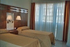 Una habitación con dos camas individuales con colchas de color beige claro y una cabecera de madera con lámparas de noche. Una ventana con cortinas claras deja pasar la luz natural.