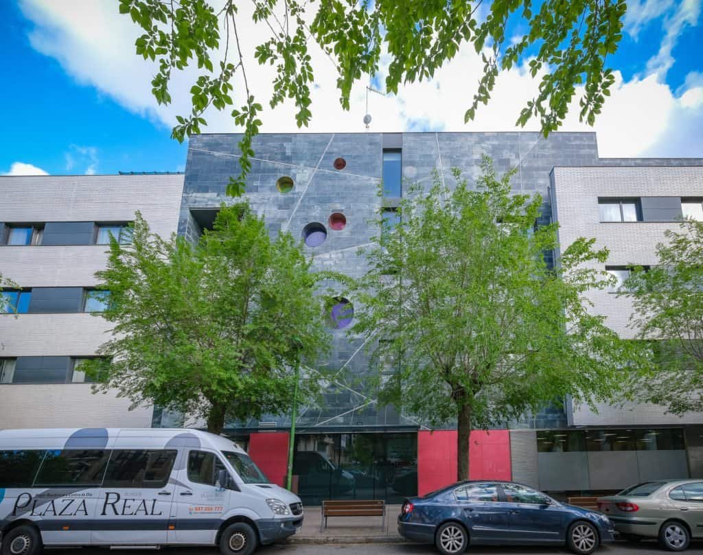 Fachada de un edificio moderno con un diseño de bloques, uno de los cuales es de piedra oscura con círculos de colores. En frente hay árboles, una furgoneta de la residencia y coches aparcados en la calle.