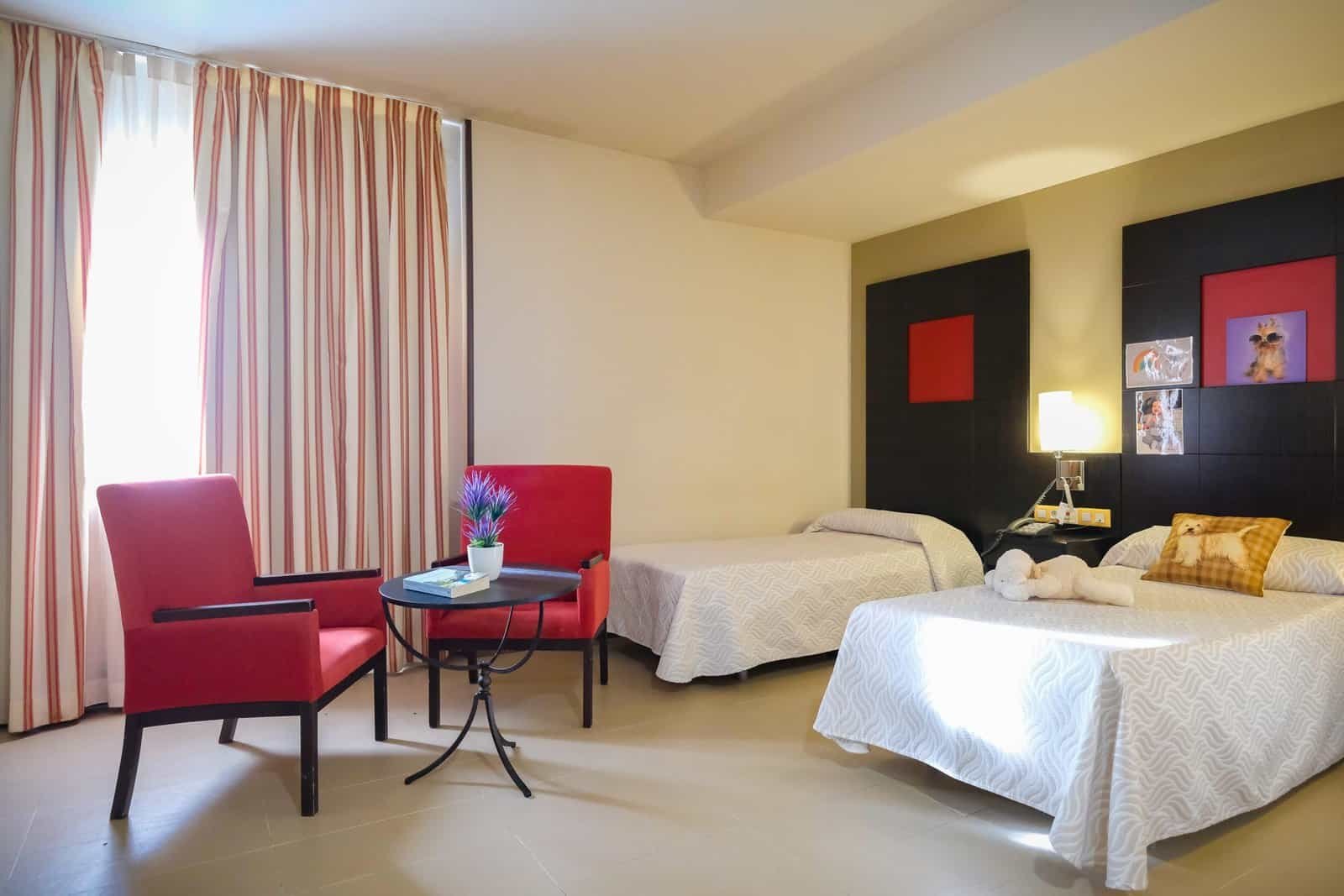Habitación doble de una residencia con dos camas, cabeceros de color oscuro con detalles rojos, dos sillones rojos y una mesa auxiliar.