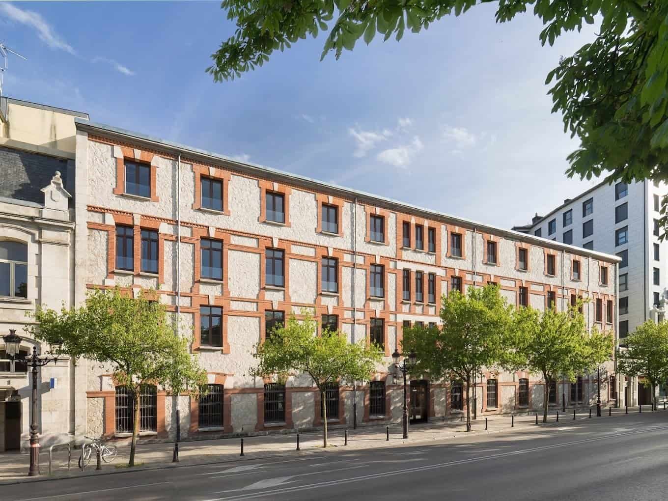 Un edificio de varios pisos con una fachada de ladrillo rojo y detalles en blanco, situado en una calle urbana arbolada con aceras y una carretera con marcas viales. El cielo está parcialmente nublado.
