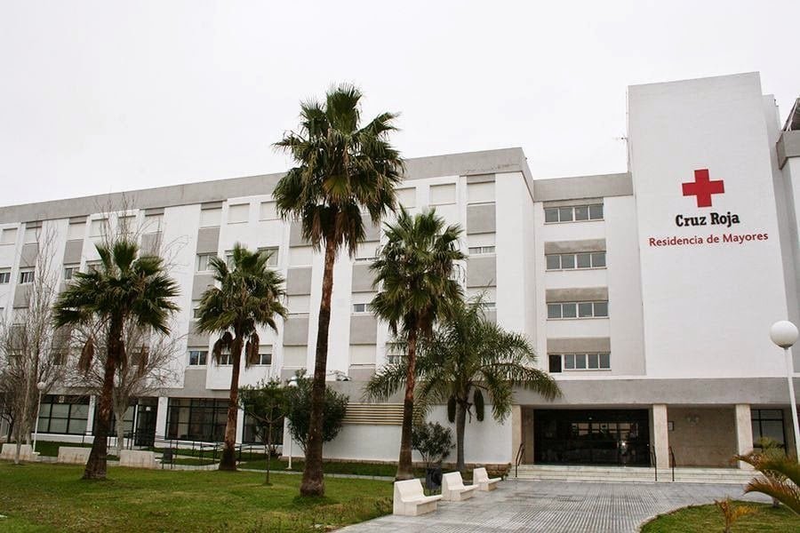 Un edificio grande y moderno, de color blanco y gris, con un gran letrero rojo de "Cruz Roja Residencia de Mayores". Hay varias palmeras en la zona ajardinada delantera.