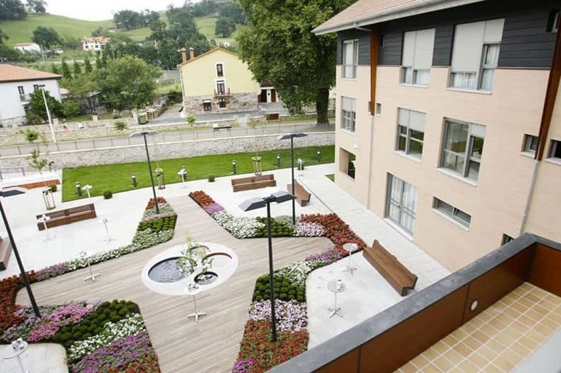 Un patio ajardinado de una residencia con pasarelas de madera, zonas de flores de colores, bancos, un pequeño estanque y un edificio en un entorno rural.