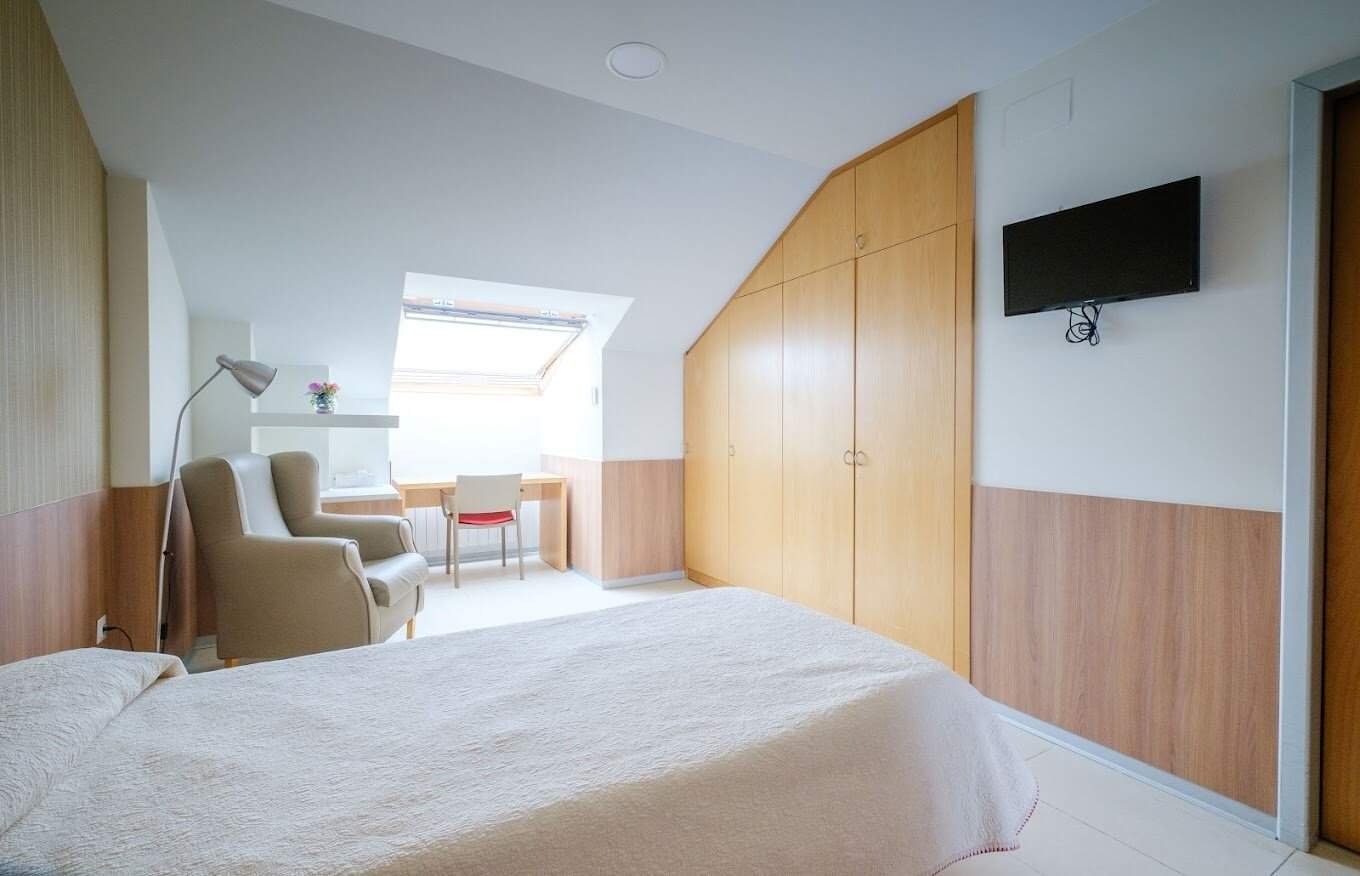 Una habitación individual moderna y luminosa en una residencia, con una cama con colcha clara, un sillón de lectura junto a una lámpara de pie, un escritorio con silla roja y un armario empotrado de madera clara. Hay una ventana de techo que aporta luz natural y una televisión plana colgada en la pared.