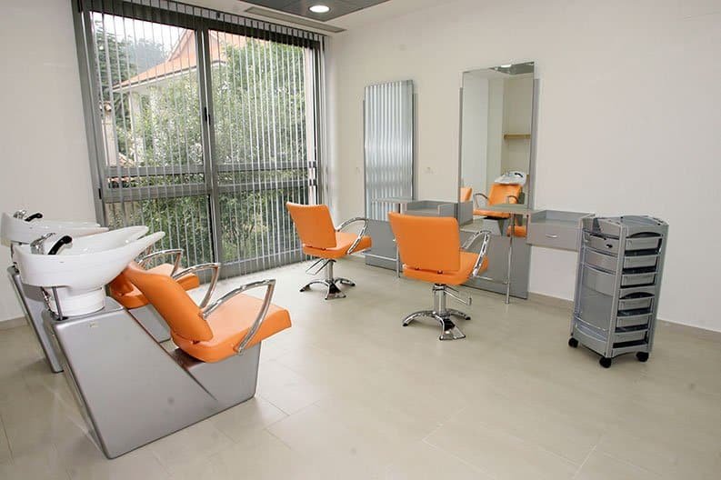 Un salón de peluquería en una residencia, con dos lavacabezas, dos sillas de peluquería de color naranja, espejos, un carrito auxiliar y un gran ventanal con persianas venecianas.