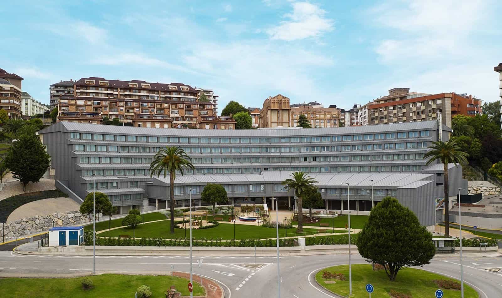 Vista exterior de un edificio de varios pisos con una fachada curvada de color gris, rodeado de palmeras y zonas verdes, en un entorno urbano.