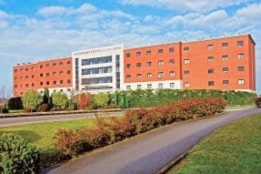 Edificio grande de ladrillo rojo con ventanas blancas. Delante hay una carretera y zonas verdes."