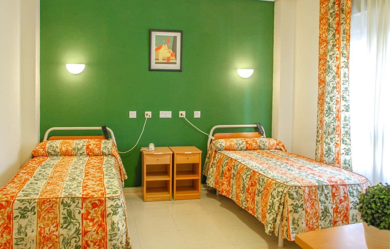 Una habitación doble con paredes de color verde brillante, dos camas individuales con colchas estampadas en tonos naranja y verde, dos mesitas de noche de madera clara y una ventana con cortinas estampadas.