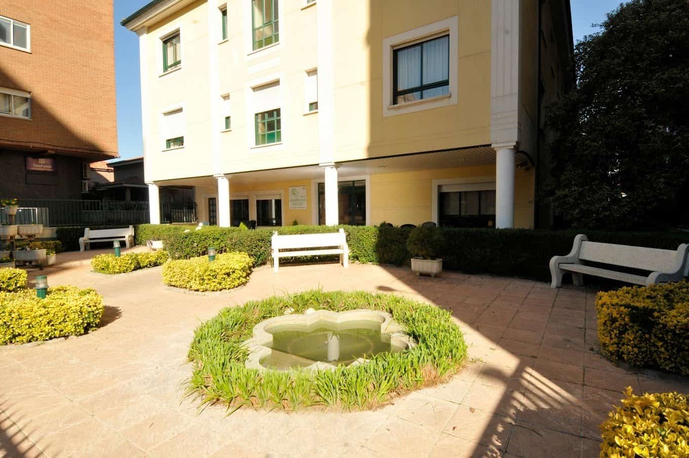 Un patio interior de una residencia con una fuente central rodeada de césped y setos. Hay bancos de color blanco y el edificio de la residencia en un segundo plano.