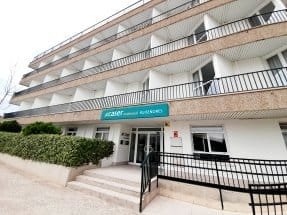 Edificio de varios pisos de la residencia Caser, con balcones y una entrada con escaleras y rampa, rodeado de una valla de setos.