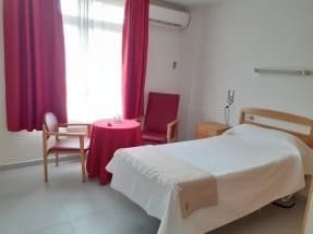 Una habitación con una cama individual con ropa de cama blanca y un sillón y una silla de color rojo, junto a una mesa pequeña redonda. Hay grandes cortinas rojas y un acondicionador de aire en la pared.