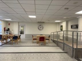 Una sala de estar o área común con mesas y sillas de madera, una rampa con barandilla metálica, un reloj grande en la pared y un suelo de baldosas con motivos decorativos.