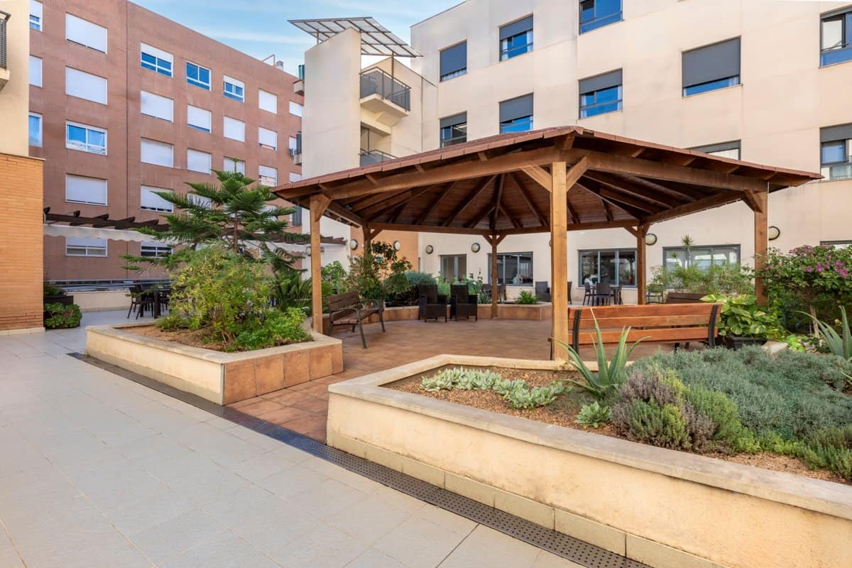 Un patio ajardinado en una residencia con un cenador de madera grande en el centro, bancos para sentarse, jardineras con plantas y árboles pequeños. En el fondo se ven edificios de varios pisos.
