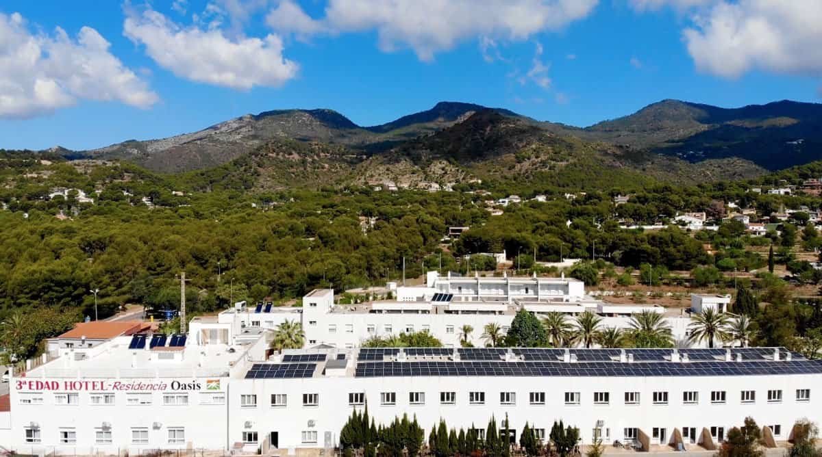 Vista aérea de un complejo de edificios blancos con paneles solares en los tejados, rodeado de vegetación y con montañas al fondo bajo un cielo azul con nubes. Se identifica como "3ª Edad Hotel - Residencia Oasis".