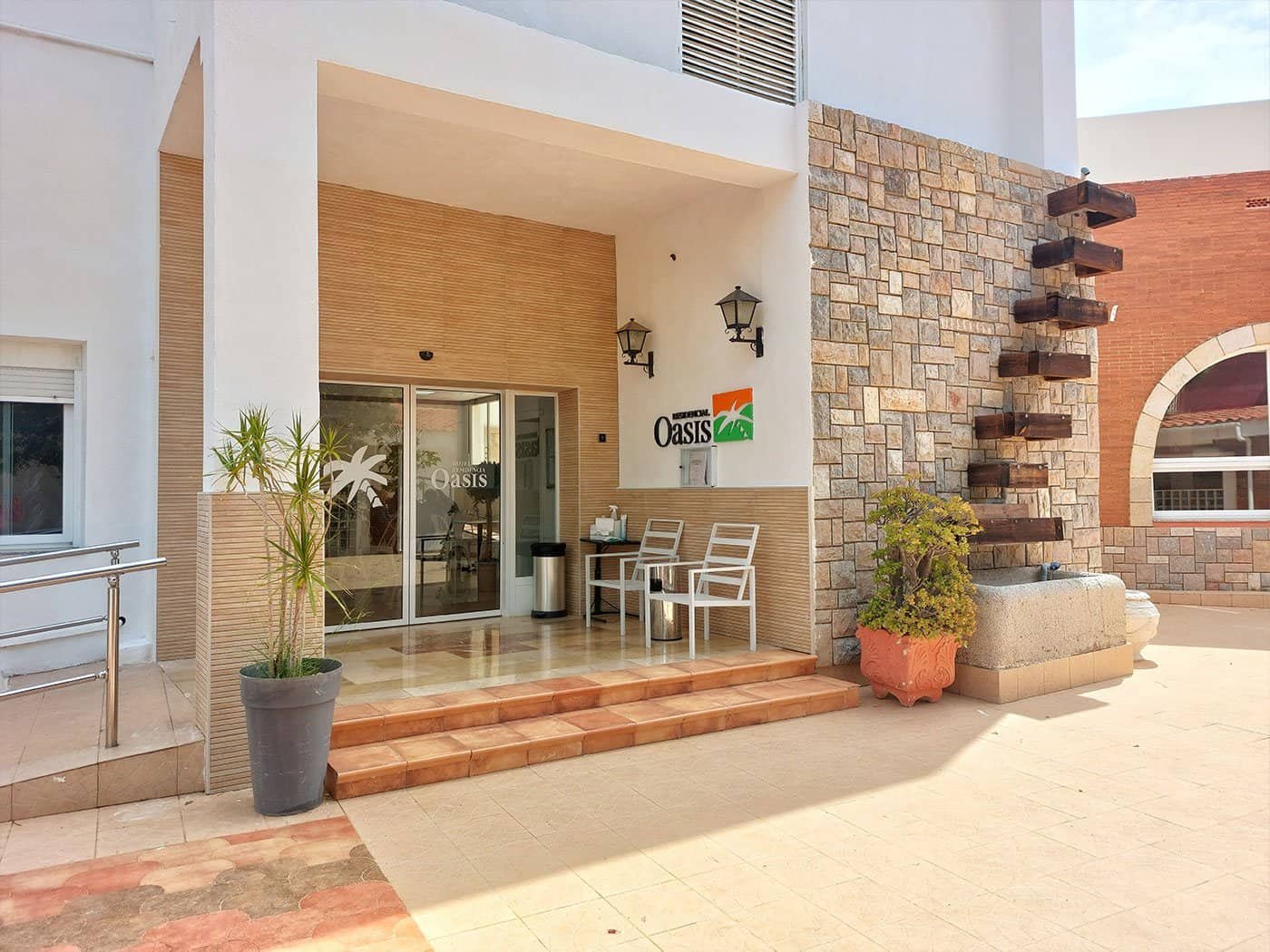 Entrada principal de la "Residencia Oasis", un edificio moderno con detalles de piedra, escalones de terracota, plantas en macetas y el logotipo del centro.