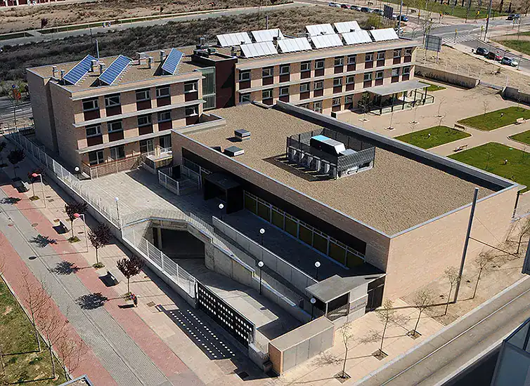 Vista aérea de un complejo de edificios de ladrillo con tejados planos, con paneles solares en la parte superior y una entrada subterránea con rampa. Alrededor hay zonas ajardinadas con árboles pequeños y aceras.