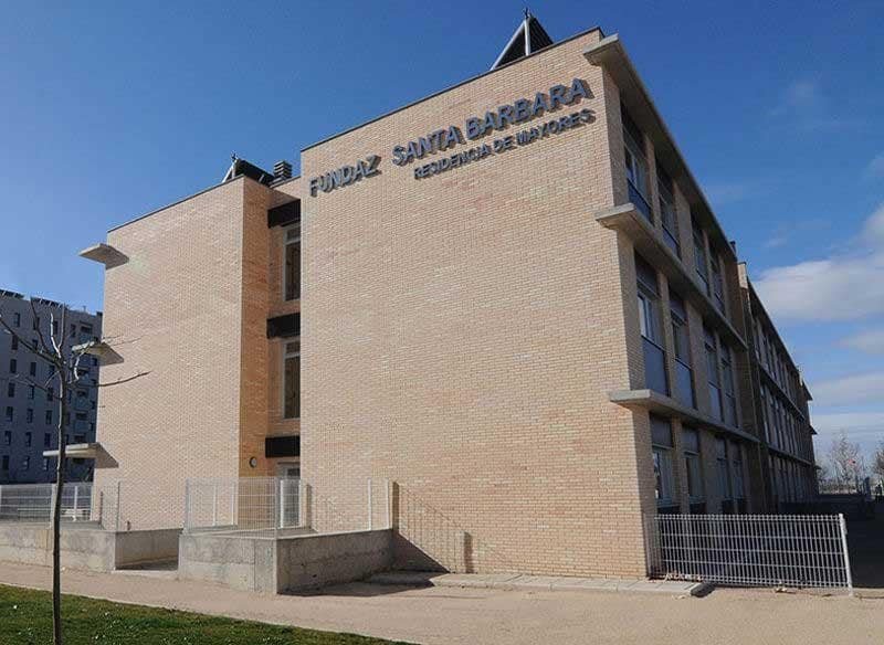 Edificio de ladrillo de varios pisos de la residencia "Fundaz Santa Barbara", con ventanas cuadradas y una valla blanca alrededor. Se ve una puerta de entrada con escaleras y un tejado a dos aguas con un panel solar.
