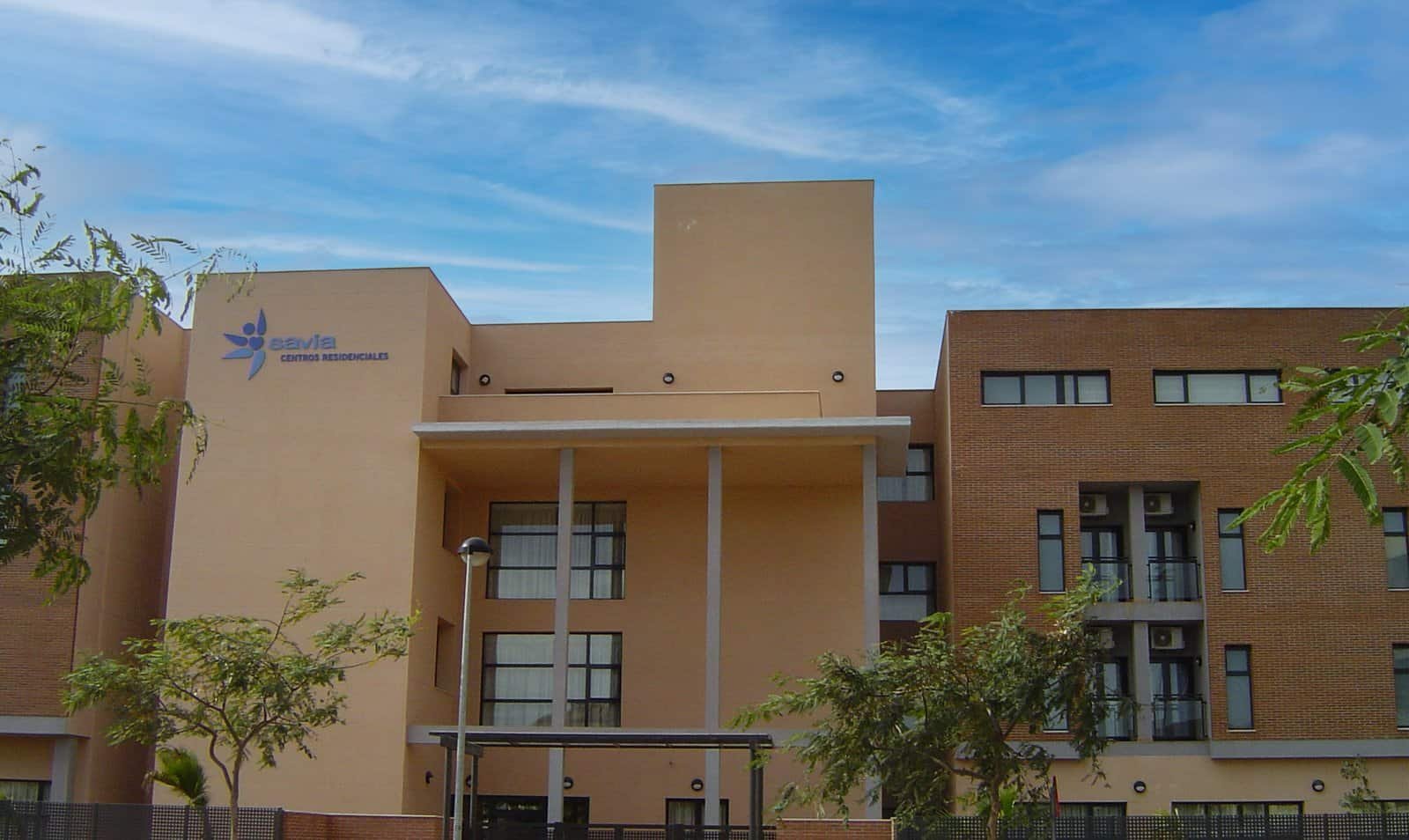 Fachada de un edificio moderno de varios pisos con diferentes secciones de color beige y ladrillo. Se aprecia un logotipo azul en la parte superior izquierda y vegetación en primer plano bajo un cielo parcialmente nublado.