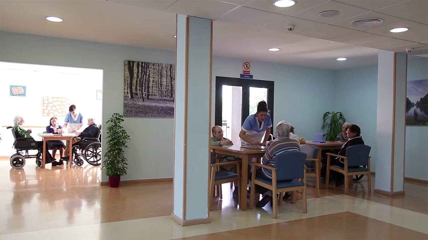 Vista interior de una residencia de ancianos con varias personas mayores realizando actividades con auxiliares. En primer plano, un grupo juega a un juego de mesa, mientras que en otra sala se ve a residentes participando en otra actividad sentados alrededor de una mesa.