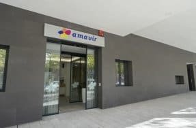 Entrada de un edificio moderno con fachada gris oscuro y el logotipo "amavir" sobre la puerta automática de cristal. Se aprecia la acera de baldosas grises y ventanas a los lados.