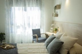 Un dormitorio luminoso con una cama con ropa de cama clara y cojines grises, un sillón azul junto a una ventana con cortinas estampadas, un escritorio de madera clara con una lámpara y decoración sencilla.