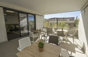 Una terraza amplia con suelo de baldosas grises y barandilla de cristal, amueblada con varias mesas y sillas de exterior de diferentes colores. Se ven edificios y árboles en el fondo.