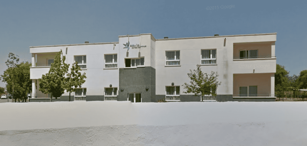 Vista frontal del edificio de la residencia Savia, de dos plantas, fachada blanca y entrada central, rodeada de árboles y en un entorno despejado.