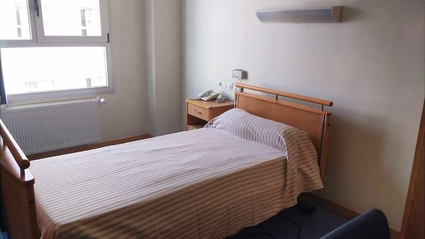 Una habitación de hospital con una cama de hospital, paredes claras, suelo de baldosas y un sillón. Se ve una ventana grande con cortinas claras.
