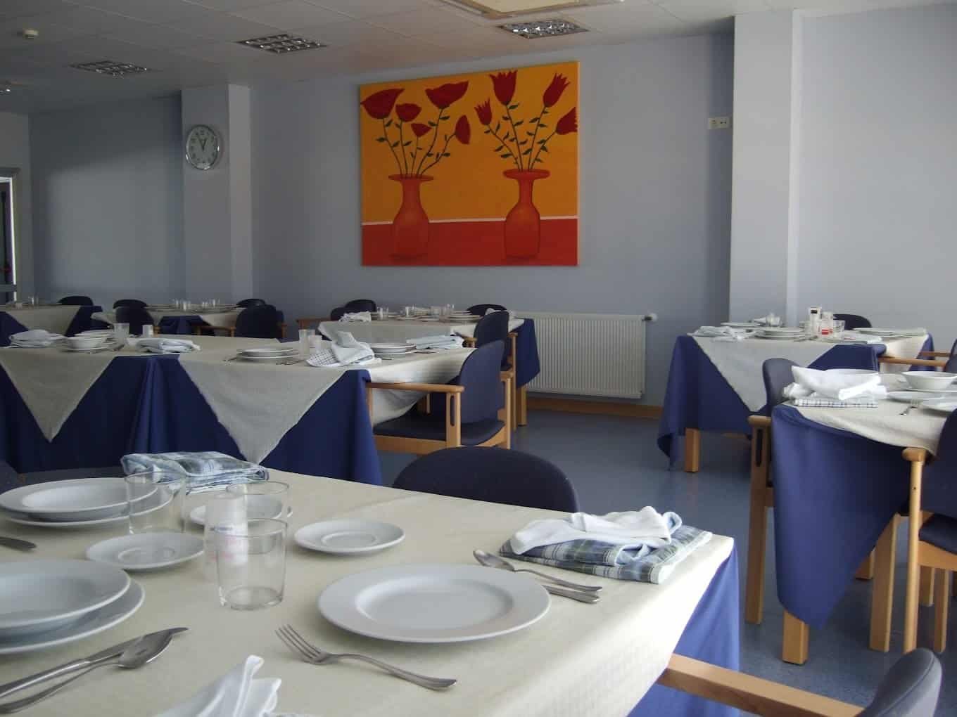 Un comedor grande y luminoso de una residencia de mayores, con mesas redondas de color claro, sillas de madera de colores, un suelo de baldosas y paredes en tonos verde claro.
