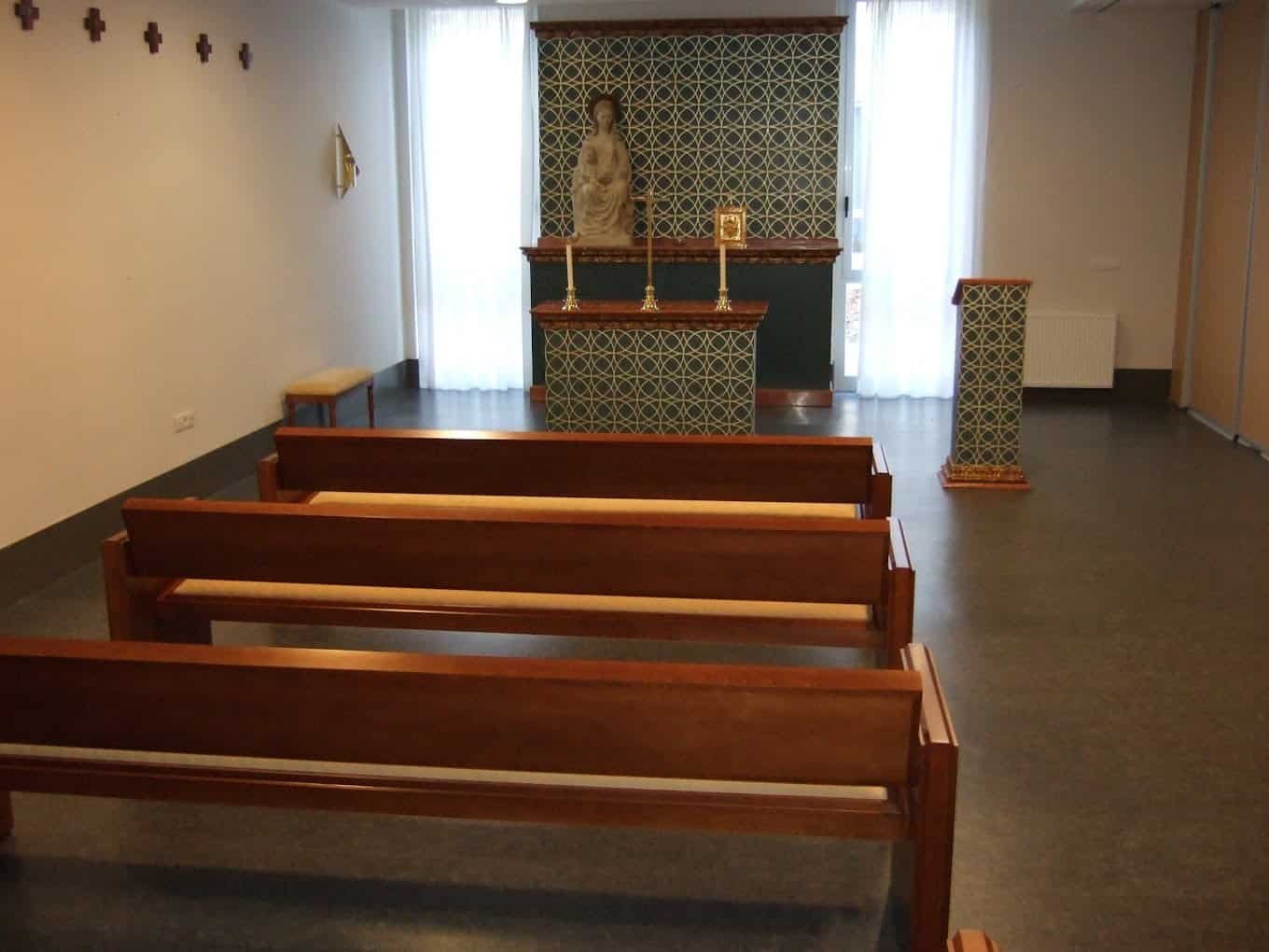 nterior de una capilla con bancos de madera, un altar al fondo y luz natural de dos ventanas laterales.