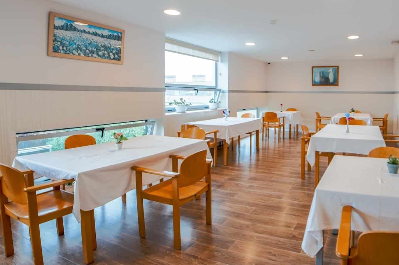 Comedor con varias mesas vestidas con manteles blancos y sillas de madera. La sala tiene ventanales y obras de arte en las paredes.