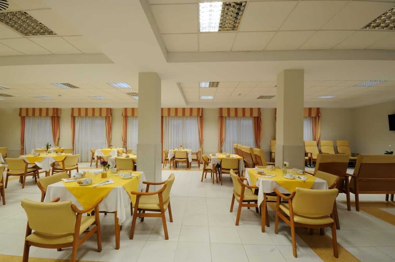 Comedor con numerosas mesas cuadradas y redondas, sillas con cojines amarillos y grandes ventanas con cortinas de rayas.
