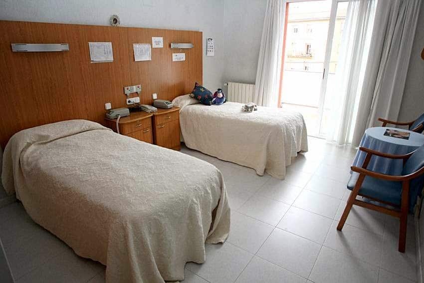 Habitación con dos camas individuales con colchas claras, una mesa y dos sillas junto a una puerta corredera que da a un balcón.