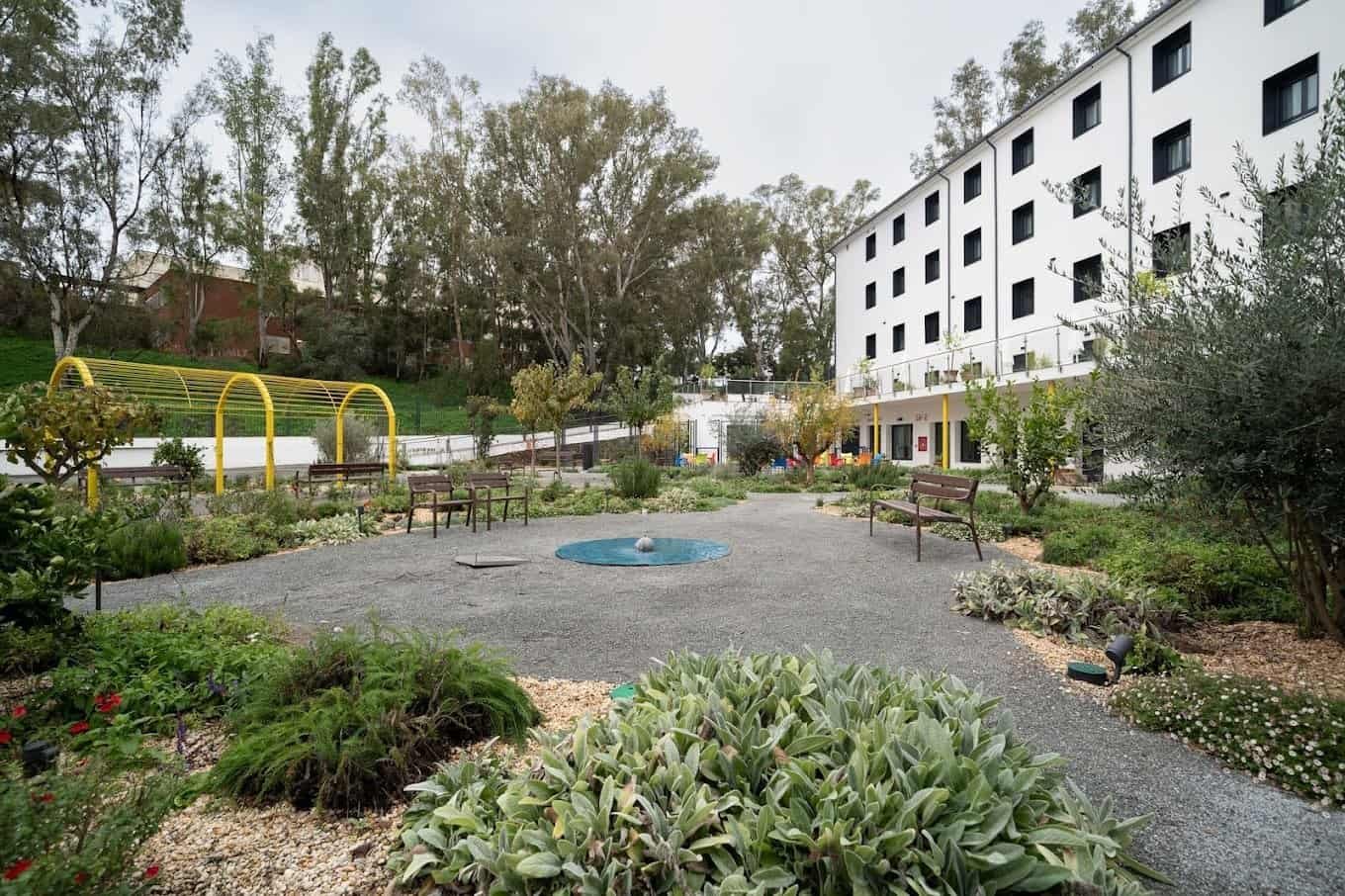 ardín exterior con zonas de gravilla y vegetación, un edificio de varias plantas al fondo y bancos de madera.