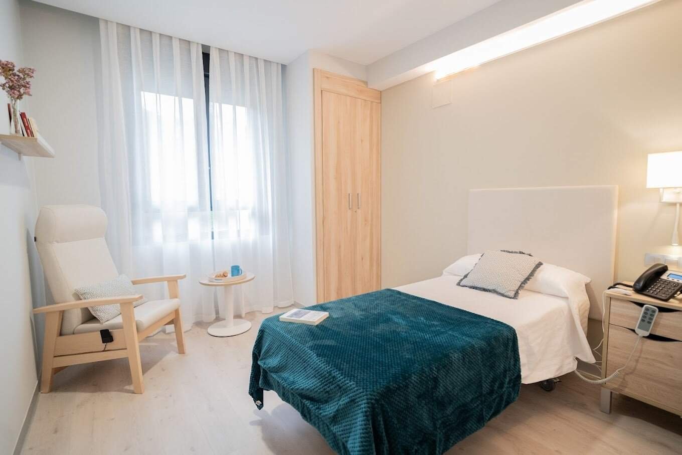 Habitación individual con cama, sillón, mesita de noche, armario de madera clara y una ventana con cortinas blancas.