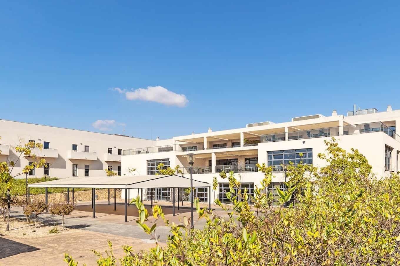 Edificio de varias plantas con una terraza grande y una zona de aparcamiento cubierta, rodeado de vegetación bajo un cielo azul.