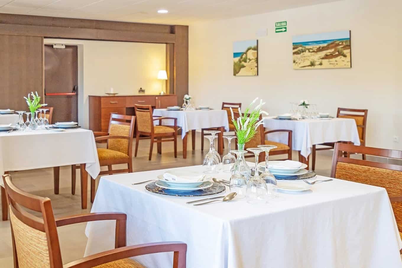 Comedor con mesas individuales puestas con vajilla, manteles blancos y jarrones de flores. Hay cuadros en la pared y un mostrador al fondo.