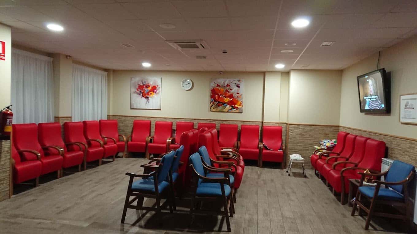 Sala común con varias filas de sillas rojas y azules, un televisor en la pared y cuadros abstractos.