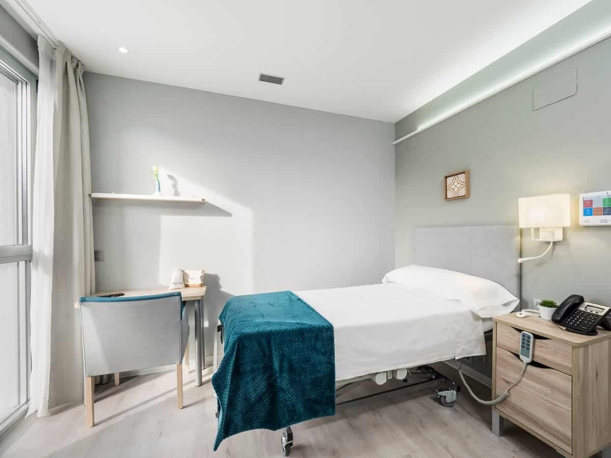 Habitación individual con una cama, un escritorio con silla y un gran ventanal con cortinas blancas.