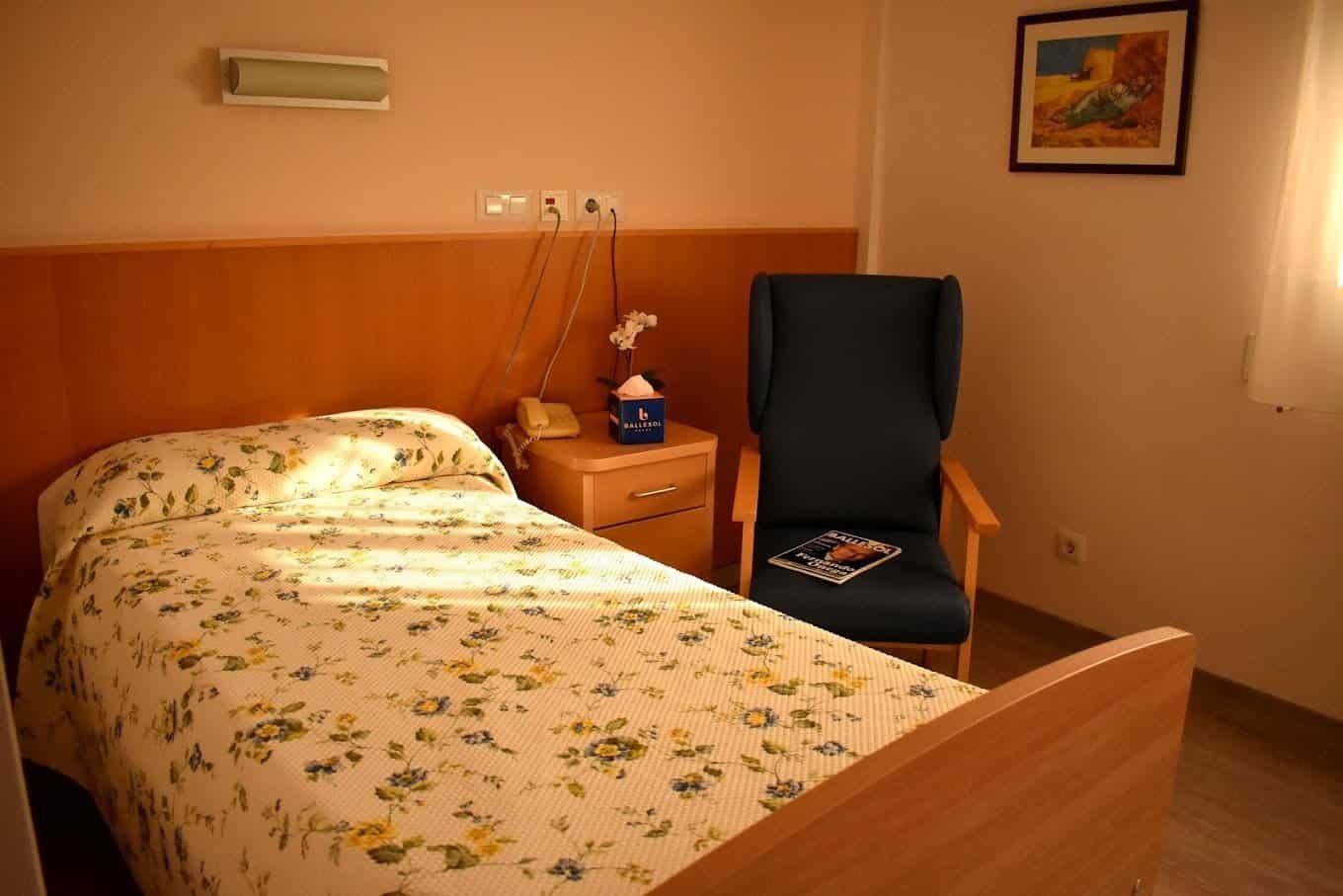 Habitación individual con una cama con colcha estampada, un sillón azul, una mesilla de noche y un cuadro en la pared.
