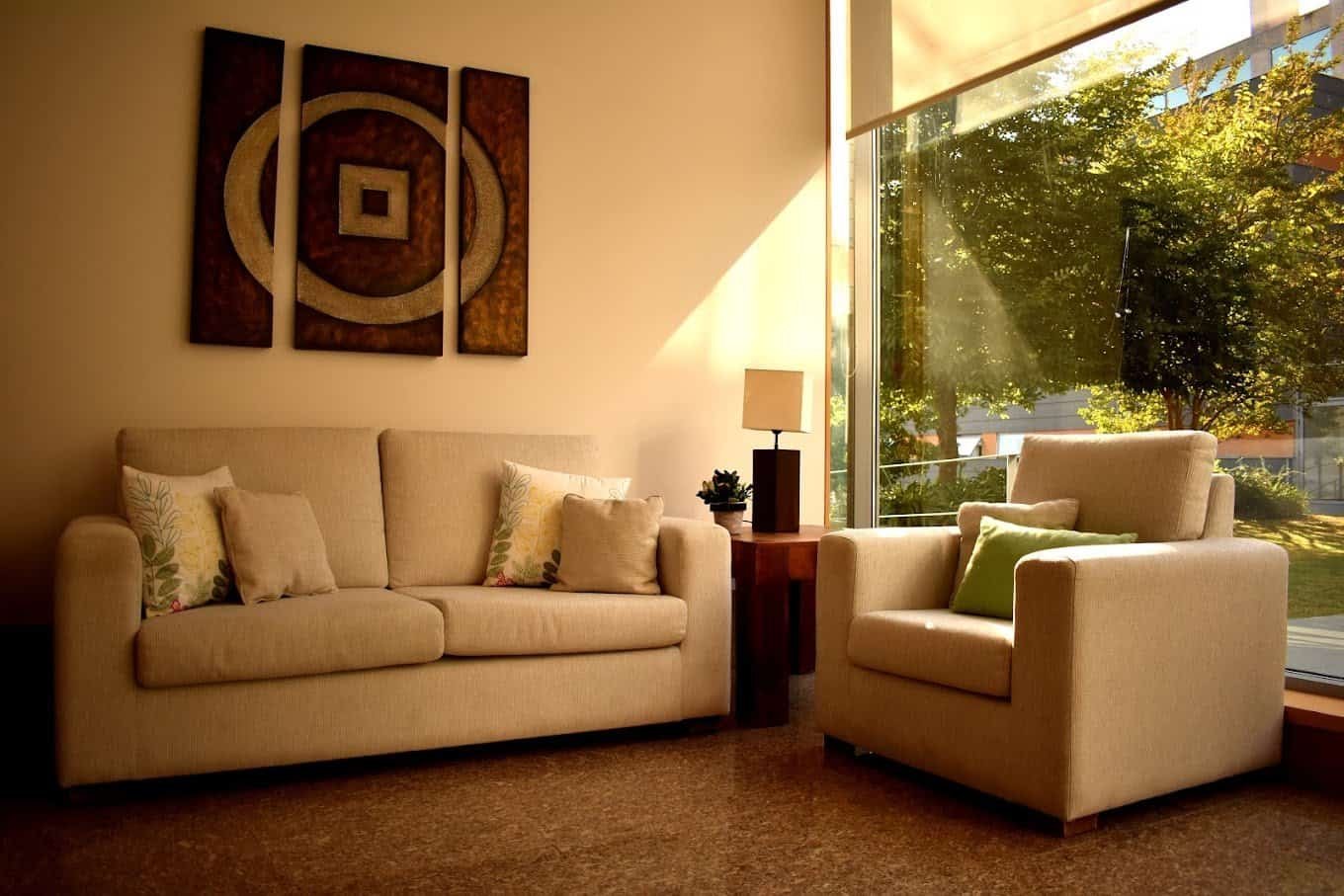 Sala de estar con un sofá y un sillón de color beige, una mesa lateral, cuadros en la pared y un gran ventanal con vistas al exterior.