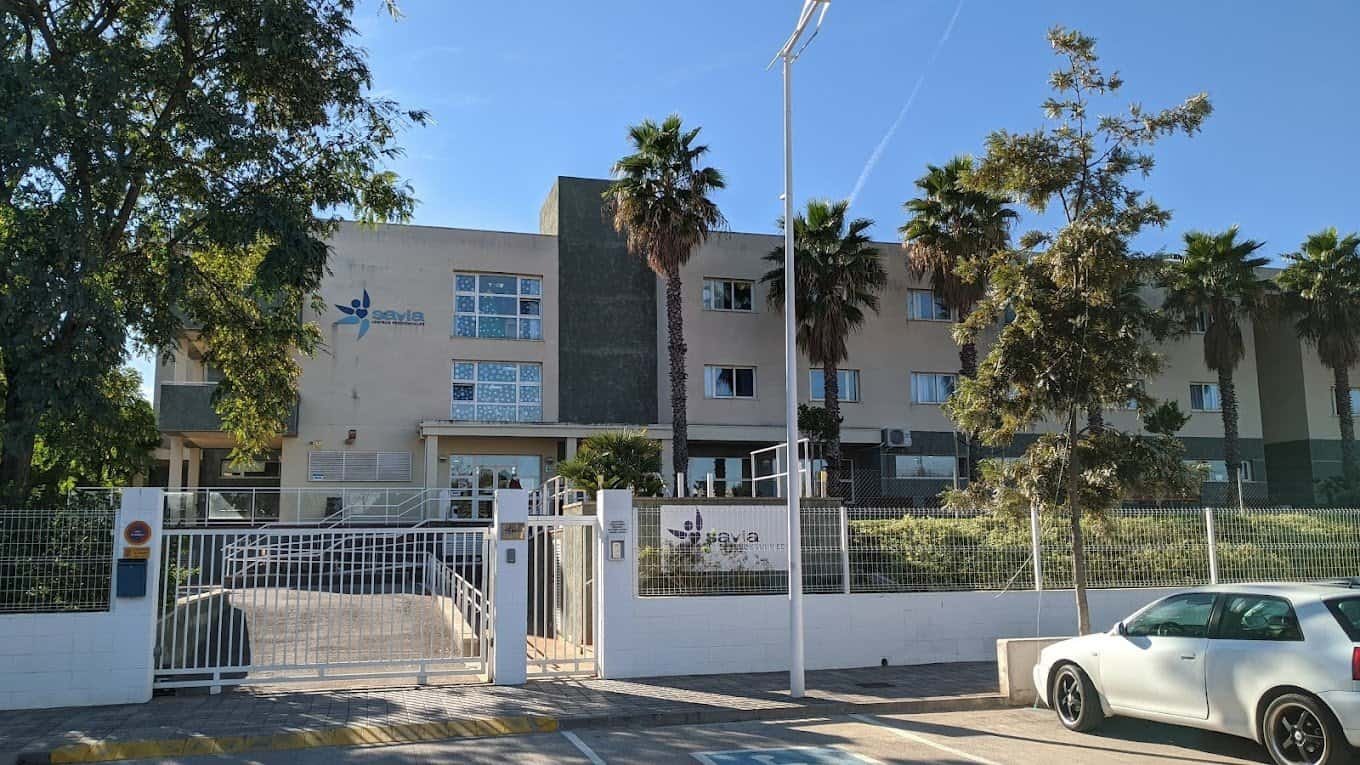 Fachada de un edificio de color crema y gris con palmeras, una valla de metal.