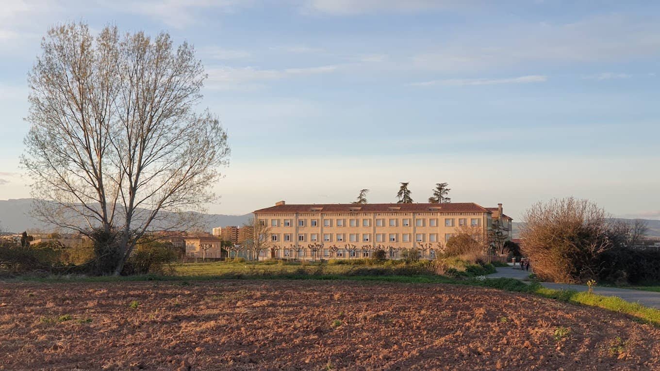 Edificio grande y antiguo de varias plantas, visto desde un campo arado con árboles y un cielo con nubes.